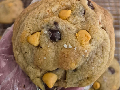 A close-up of a large chocolate chip cookie butterscotch morsels,chocolate chips, and pretzels with coarse sea salt sprinkled on top.