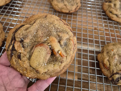 A hand holding a large chocolate chip cookie with visible chocolate chunks, shown over a metal cooling rack with more cookies on a wooden surface.