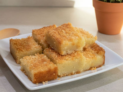 Six square slices of golden-brown coconut cake bars stacked on a white square plate