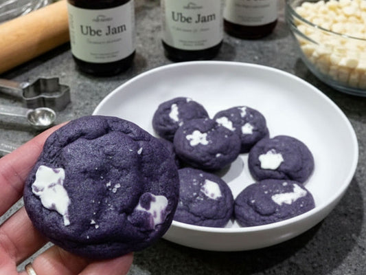 A hand holds a purple ube jam cookie in the foreground, with several more cookies in a white bowl behind it. Ube jam bottles and baking ingredients are visible in the background.