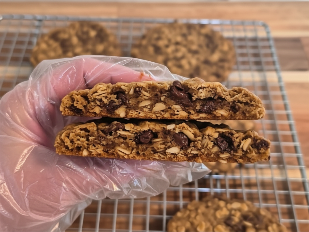 A hand wearing a clear plastic glove holds two halves of a sliced granola cookie, showing oats, chocolate pieces, More cookies rest on a metal cooling rack in the background.