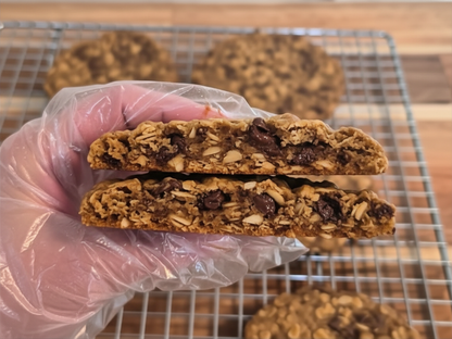 A hand wearing a clear plastic glove holds two halves of a sliced granola cookie, showing oats, chocolate pieces, More cookies rest on a metal cooling rack in the background.