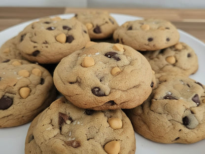 A pile of salty butterscotch chocolate chip cookies with pretzels on a white plate, photographed close-up on a light wooden surface.
