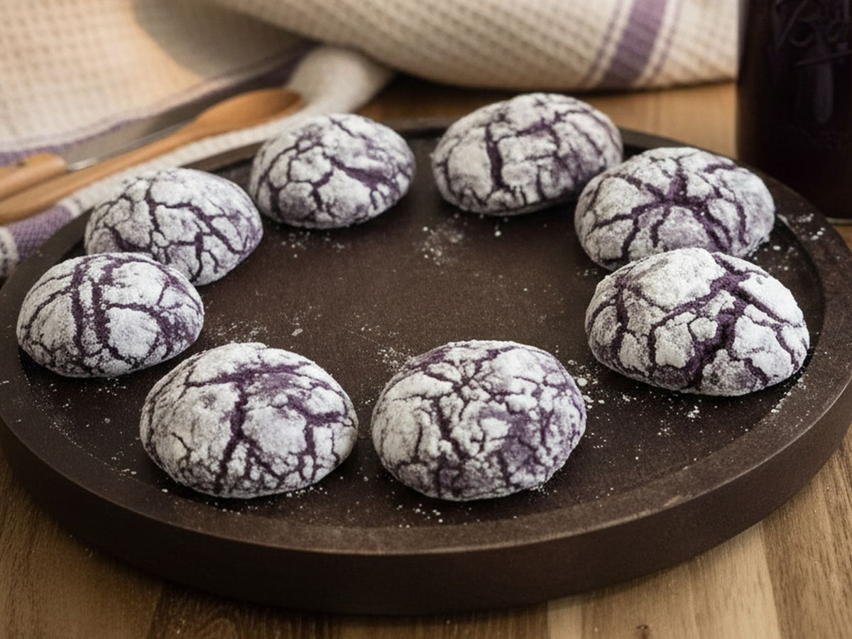 Eight ube mochi purple crinkle cookies dusted with powdered sugar, arranged in a circle on a dark round wooden serving board over a wooden table.